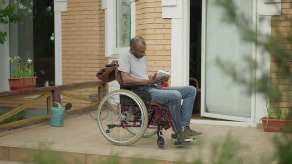 Absorbed Handicap African American Man Reading Book Sitting in Wheelchair on Porch Outdoors alt