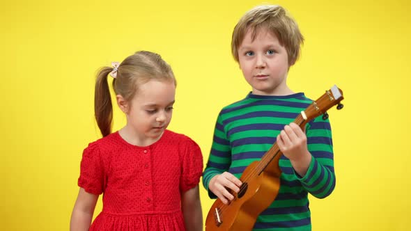 Charming Caucasian Brother and Sister Playing Ukulele at Yellow Background alt