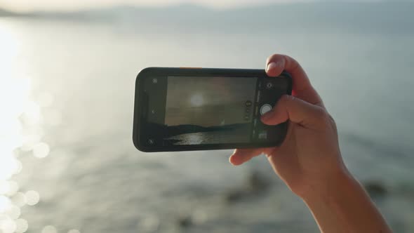 Closeup of a Phone in Female Hands Making a Photo at Sunset By the Sea alt