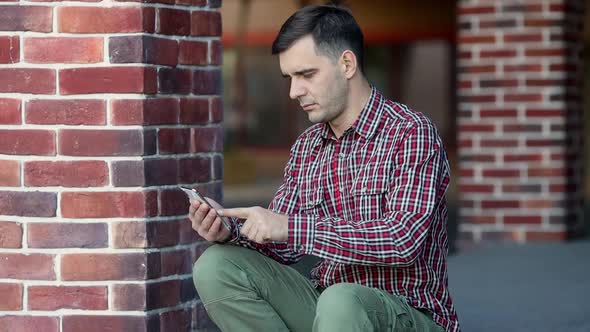 Urban portrait of serious handsome millennial man using mobile phone alt