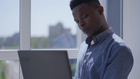 Young Concentrated Man Thinking Surfing Internet on Laptop Standing at Window in Home Office alt