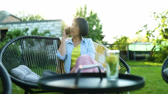 Happy Woman in Glasses Talking on Mobile Phone Sitting on Chair at Backyard Outdoor
