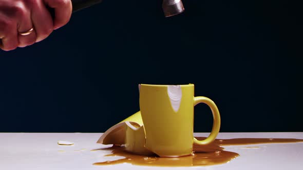 Man's Hand Breaks a Yellow Tea Mug with Liquid with an Iron Hammer
