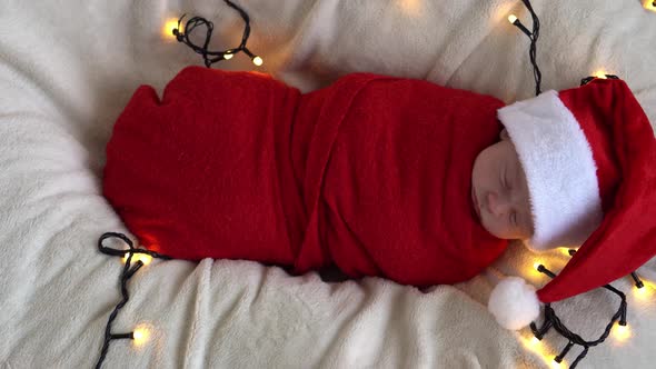 Top View Portrait First Days Of Life Newborn Cute Funny Sleeping Baby In Santa Hat Wrapped In Red