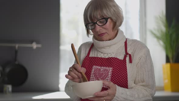 Medium Shot Portrait of Positive Senior Woman Mixing Smelling Sauce in Bowl Smiling alt