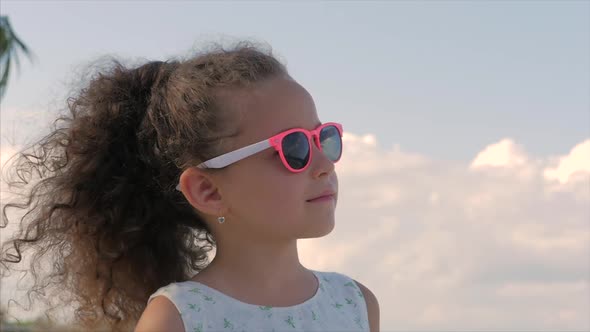 Close-up Portrait of a Beautiful Little Girl in Pink Glasses, Cute Smiling, Looking at the Blue Sky alt