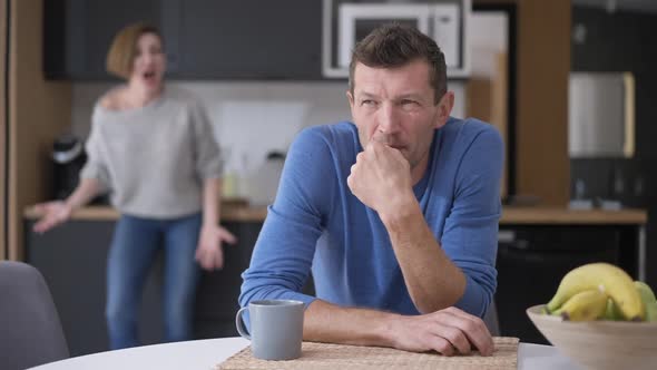 Sad Stressed Bored Man Sitting at Table Sighing As Woman Yelling at Background Arguing alt