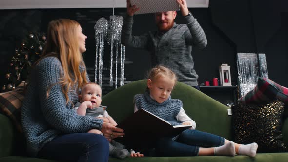 New Year - Mom with Children Is Sitting on the Couch and Reading Books and Dad Is Coming with Gifts alt