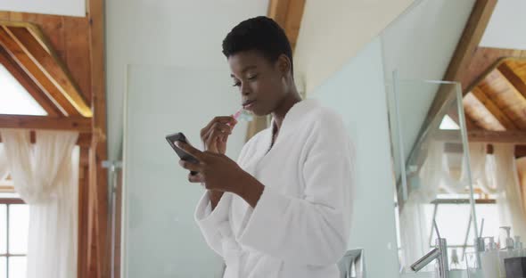 African american attractive woman brushing teeth and using smartphone in bathroom alt