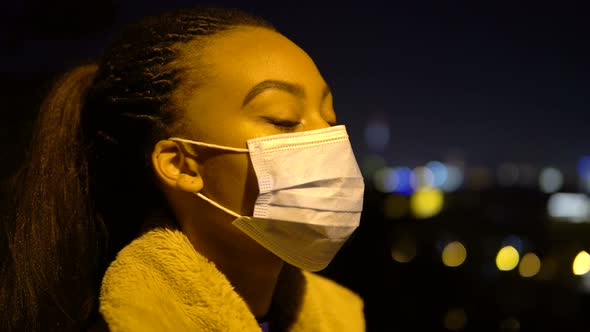 A Young Black Woman in a Face Mask Looks Around in an Urban Area at Night  Side Closeup alt