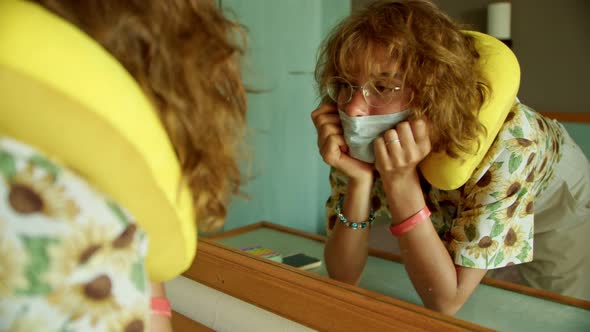 a Tourist is Looking at Herself in the Mirror in a Hotel Room and Fixing a Medical Protective Face alt
