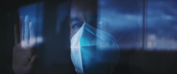 Man in a face mask looking out of a window during the isolation alt