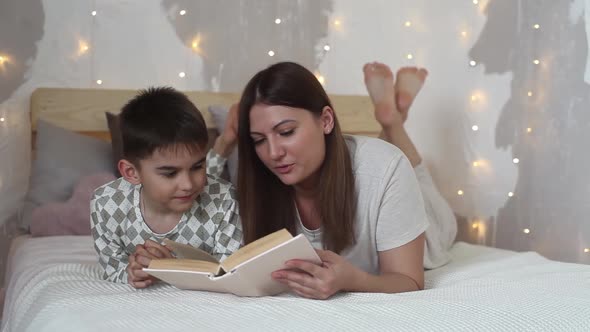 Beautiful Girl with Long Hair Reads a Book to a Small Boy Lying on a Bed and Kisses Him alt