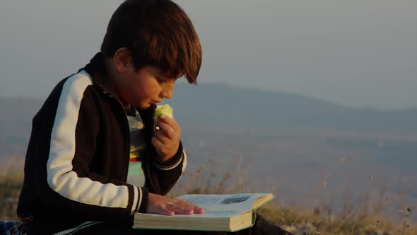 Boy Reads a Book and Eats an Apple in the Rays of the Sunset on the Background of Mountains