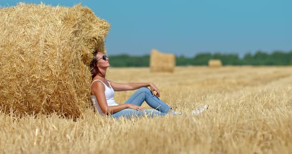Beautiful Woman in Sunglasses is Sitting By a Large Haystack She is Resting in a Field alt