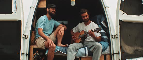 Two friends sitting in back of a van having fun and playing the guitar alt