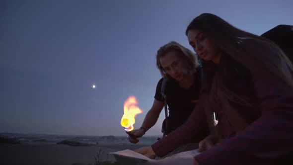 Young Couple Using a Flaming Torch To Read a Map at Dusk in the Wilderness During a Hiking Trip with alt