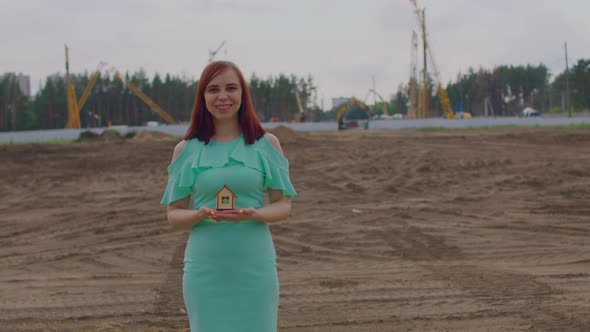 Young Woman with Small Wooden House in Hands Stands on Construction Site alt
