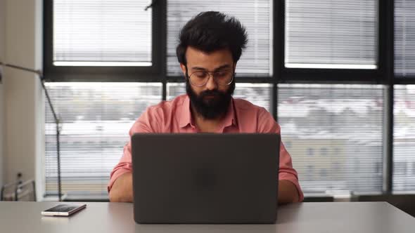 Front View of Thoughtful Bearded Young Business Man Typing on Laptop Computer Sitting at Desk in alt