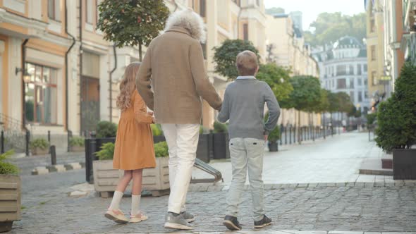 Wide Shot of Grandfather and Grandchildren Walking Along Paving Street on Sunny Day. Back View of alt