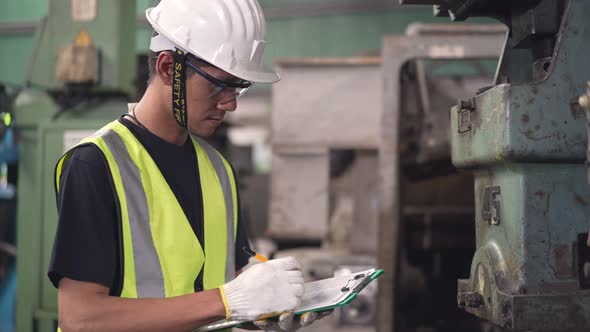 Asian male engineer or factory worker in safety clothing writing on checklist clipboard alt
