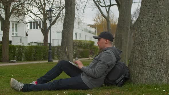 Young Man is Sitting in a Park in a Tourist Place Working on a Laptop alt