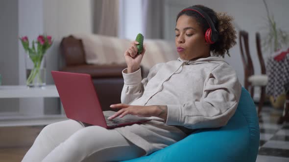 Concentrated African American Woman Eating Cucumber Typing on Laptop Keyboard Sitting on Bag Chair alt
