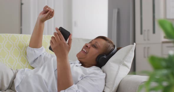 Happy african american senior woman wearing headphone lying on couch using smartphone and smiling alt