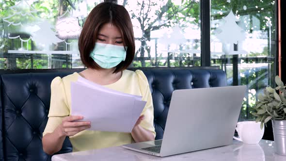 Young Asian businesswoman wearing masks and checking documents on paper and laptop alt