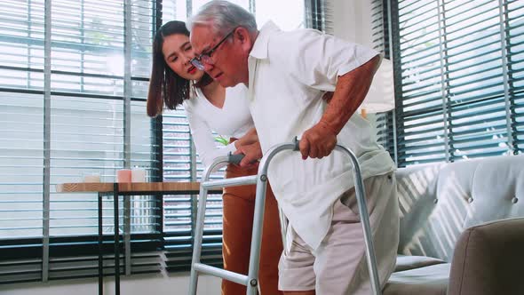 Daughter is helping the disabled father learn to walk with walking frame in the house. alt