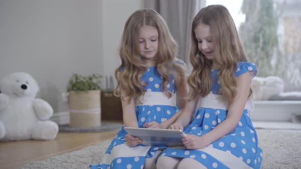 Portrait of Two Identical Twin Sisters in Similar Dresses Using Tablet at Home. Little Caucasian alt
