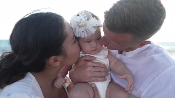 Pretty Baby Girl at Mediterranean Sea Beach with Father and Mother Kissing Daughter on Cheeks alt