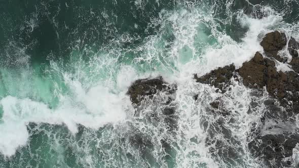 Aerial top down view of waves splashing into rocks in the sea, super slow mot