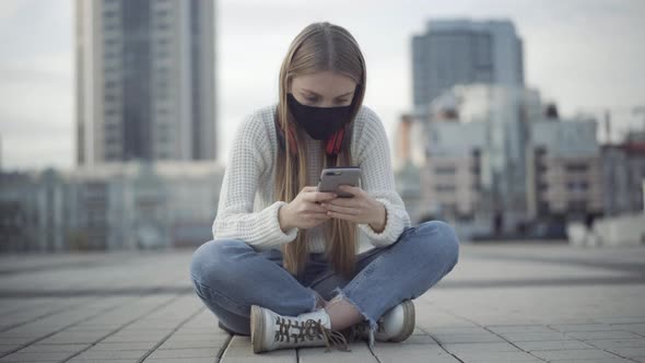 Portrait of Concentrated Young Woman in Covid Face Mask Sitting on City Square and Messaging Online alt