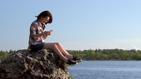 A Young Woman Sits on a Stone By the River with a Phone in Her Hands. The Girl Is Texting in the alt
