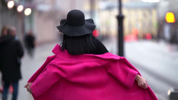 Fashionable Glamorous Young Woman with Black Hair, Black Hat and Purple Coat Walks Down the Street alt