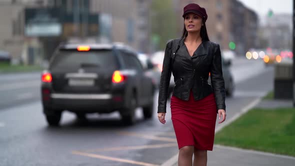 a Brunette in a Black Leather Jacket a Red Skirt and a Burgundy Cap Walks Down the Street Against alt