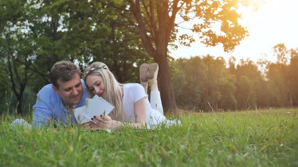 A Guy and a Girl are Watching a Family Album with Photos in the Park on the Grass