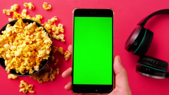 Man's hand holding smart phone with green touch screen and popcorn in a bowl on red background. alt