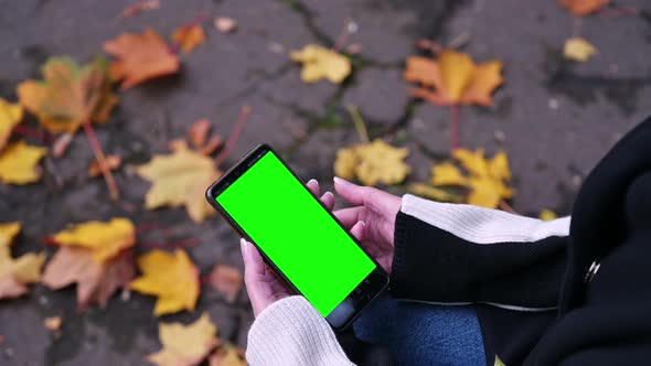 A woman's hand holds a mobile phone with a green screen while sitting in the park on a bench