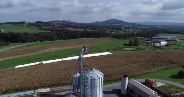 Aerial view pulling away from American flag at top of grain silo with cornfields and mountains in th alt