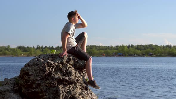 A Young Man Sits on a Stone By the River and Admires Nature. A Man in Nature Closeup. alt