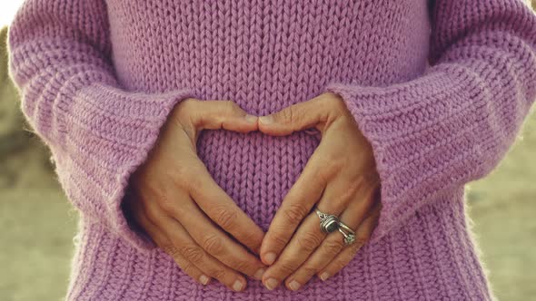 Woman hands do hearth sign on belly against violet sweater. Romantic and newborn life pregnant alt