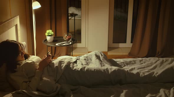 Girl in Glasses Lies in Bed at Night Covered with a Blanket Uses a Mobile App Camera Tracking alt