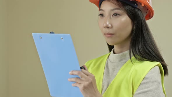 Asian Builder Woman Wearing Protective Clothing and a Helmet is Writing Down Something alt