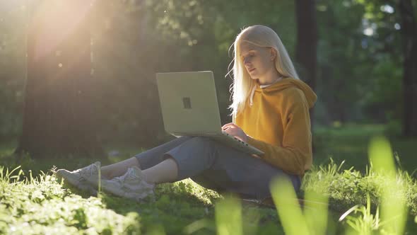 Beautiful Blonde Woman Sitting on Grass Under a Tree at Park and Working on Laptop