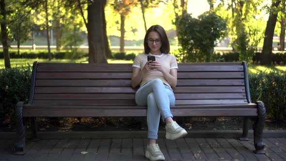 A Young Woman in Glasses Sits on a Bench with a Phone in Her Hands. In the Background Nature alt