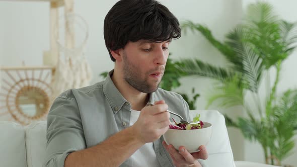 Young Brunette Man Eating Fresh Vegetable Salad While Sitting on the Couch at Home alt