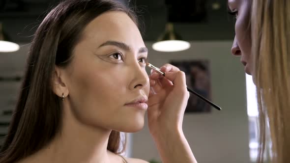 Make Up Artist Doing Makeup for Brunette Asian Woman with Brush in Beauty Salon