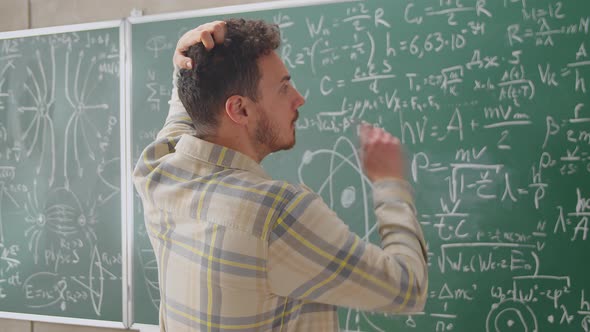 Young Man is Walking to Blackboard Wall in College Class Looking at Formulas Thinking About Science alt